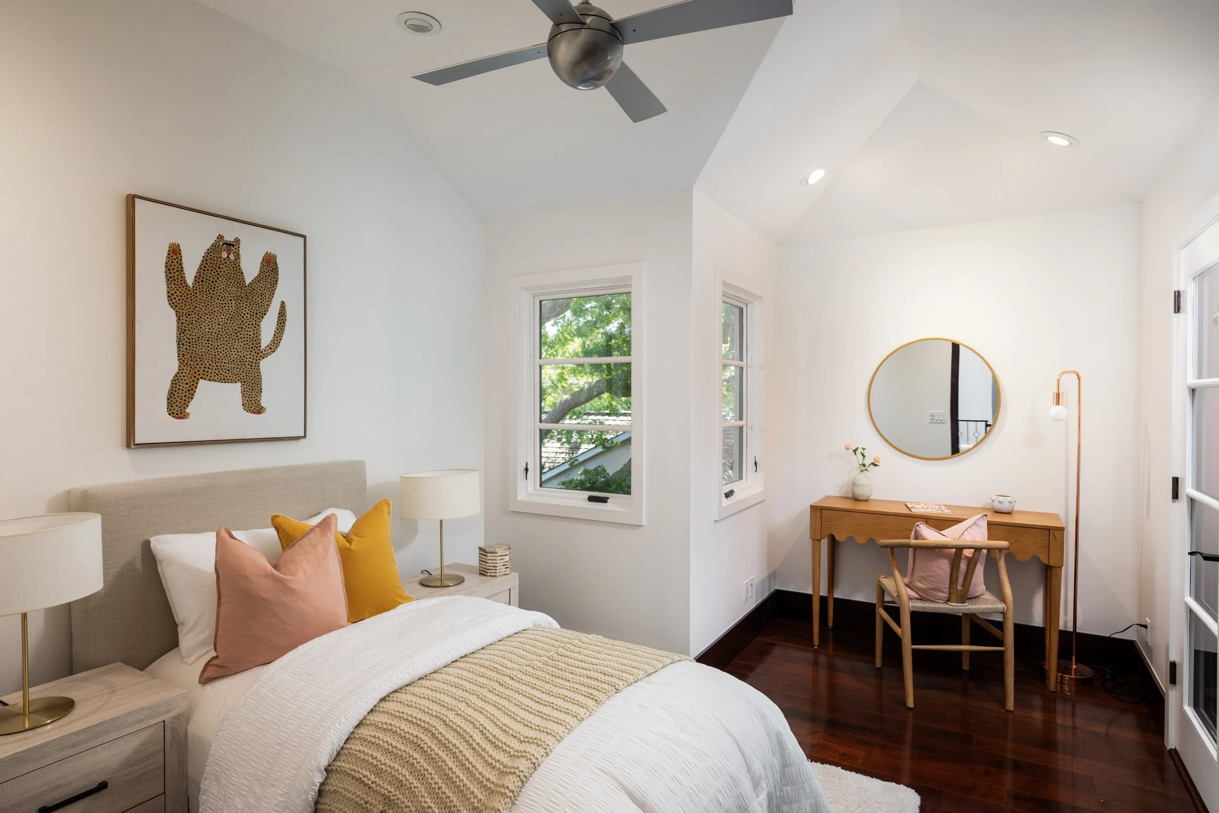 A bedroom with a beige bed, decorative pillows in pink and yellow, white nightstands with lamps, artwork of a cheetah on the wall, three windows showing green trees outside, a round mirror, a wooden desk, a wooden chair with pink cushions, a standing
