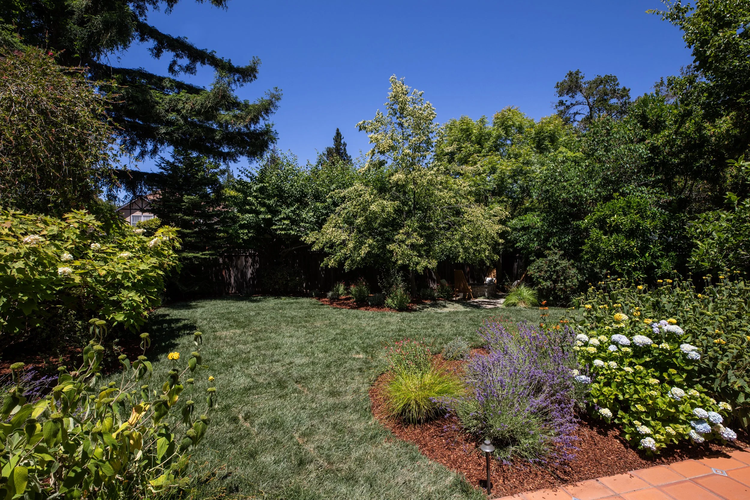 A lush backyard garden under a clear blue sky features a well-maintained lawn, colorful flower beds, and tall green trees providing shade.
