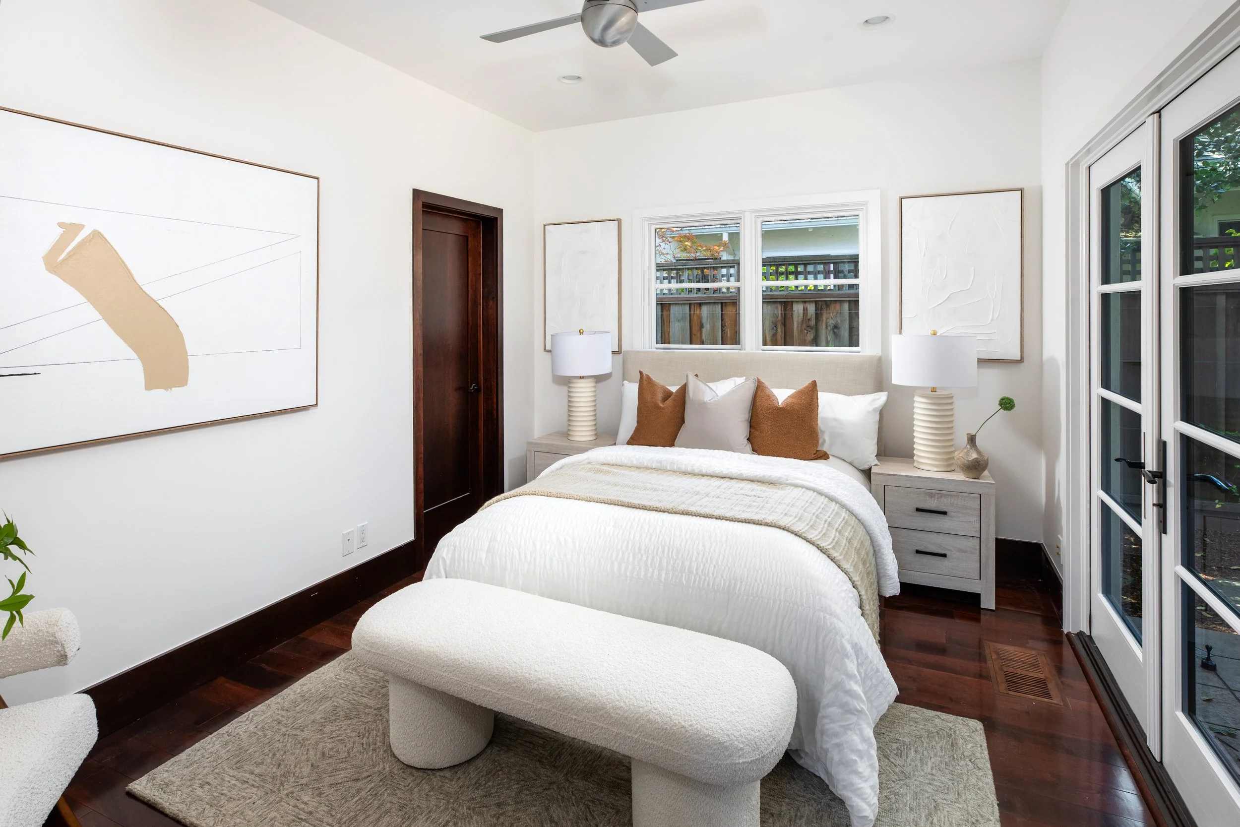 A bedroom with a white bed, beige headboard, white bedding, brown and white pillows, and nightstands with white lamps. There is abstract artwork on the walls, a window showing an outside wooden fence, and a ceiling fan.