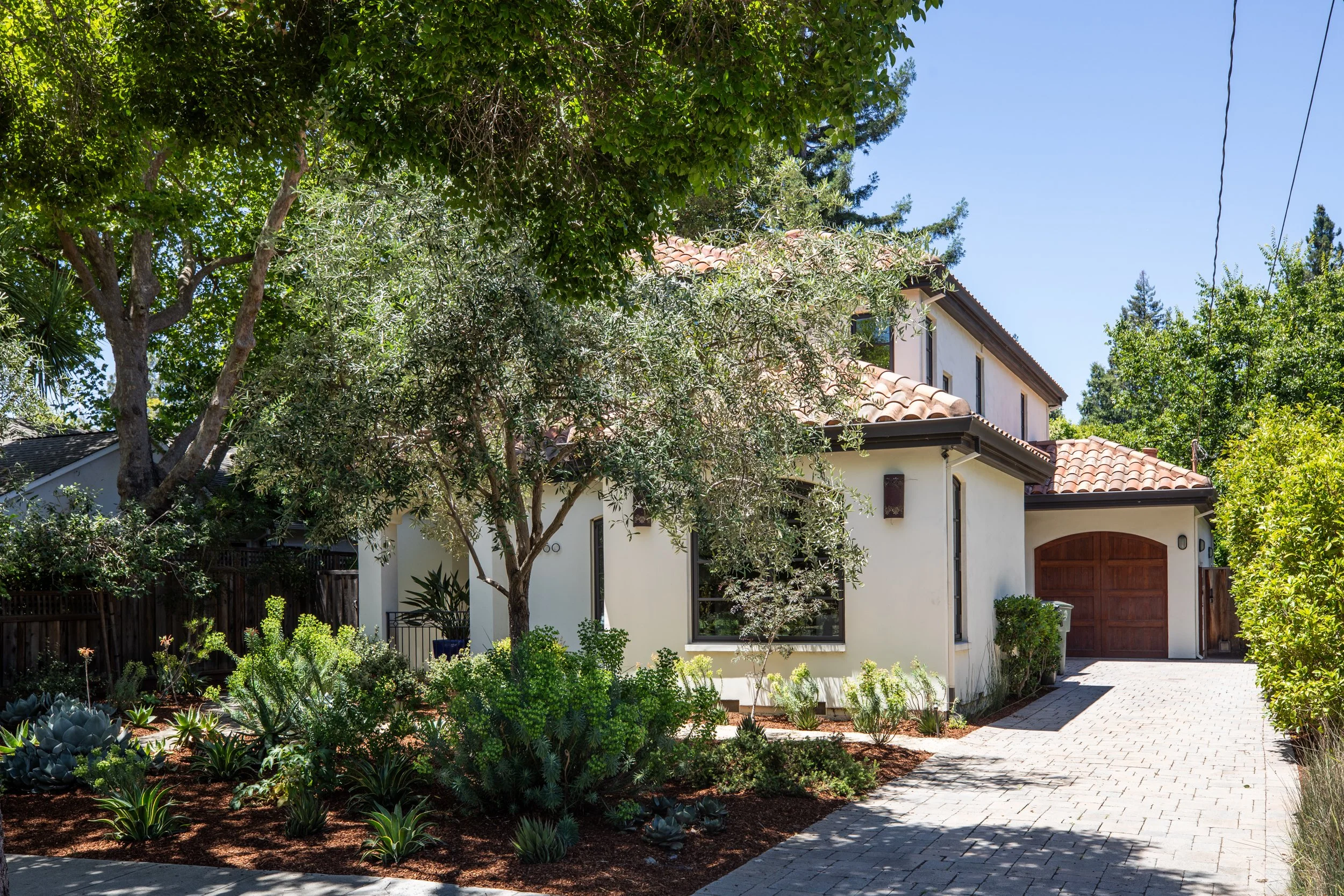 A house with a Spanish tile roof surrounded by lush green trees and a garden with succulents, under a clear blue sky.