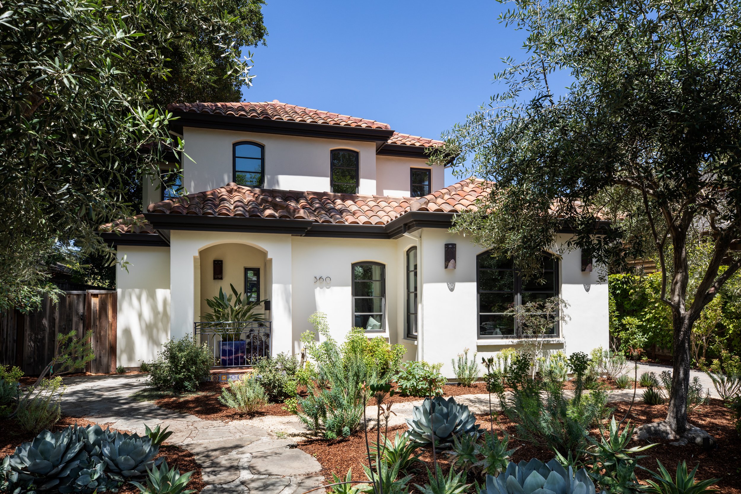 White two-story house with a terracotta tile roof, surrounded by lush green plants and trees, under a clear blue sky.