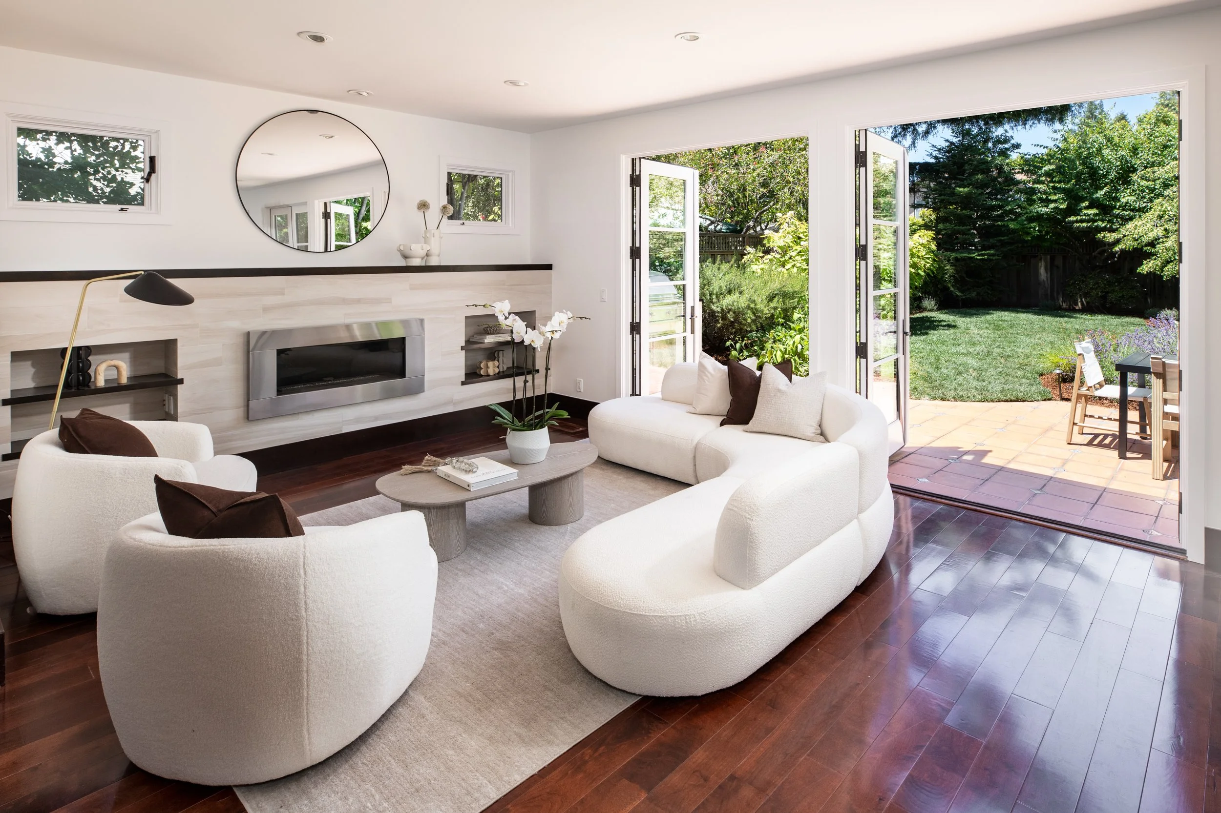 Living room with white couches, dark wood floors, a fireplace, large glass doors open to a backyard with green trees and a patio.