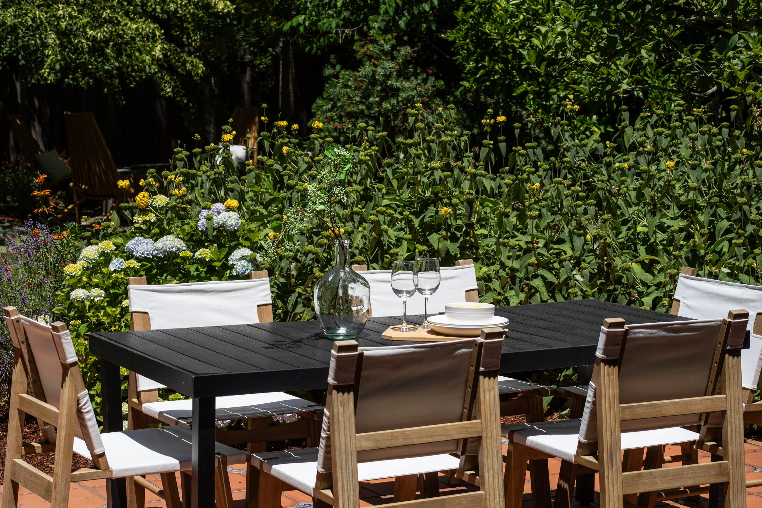 Outdoor dining table with white chairs, glassware, and bowls, set in a lush green garden with flowering plants.
