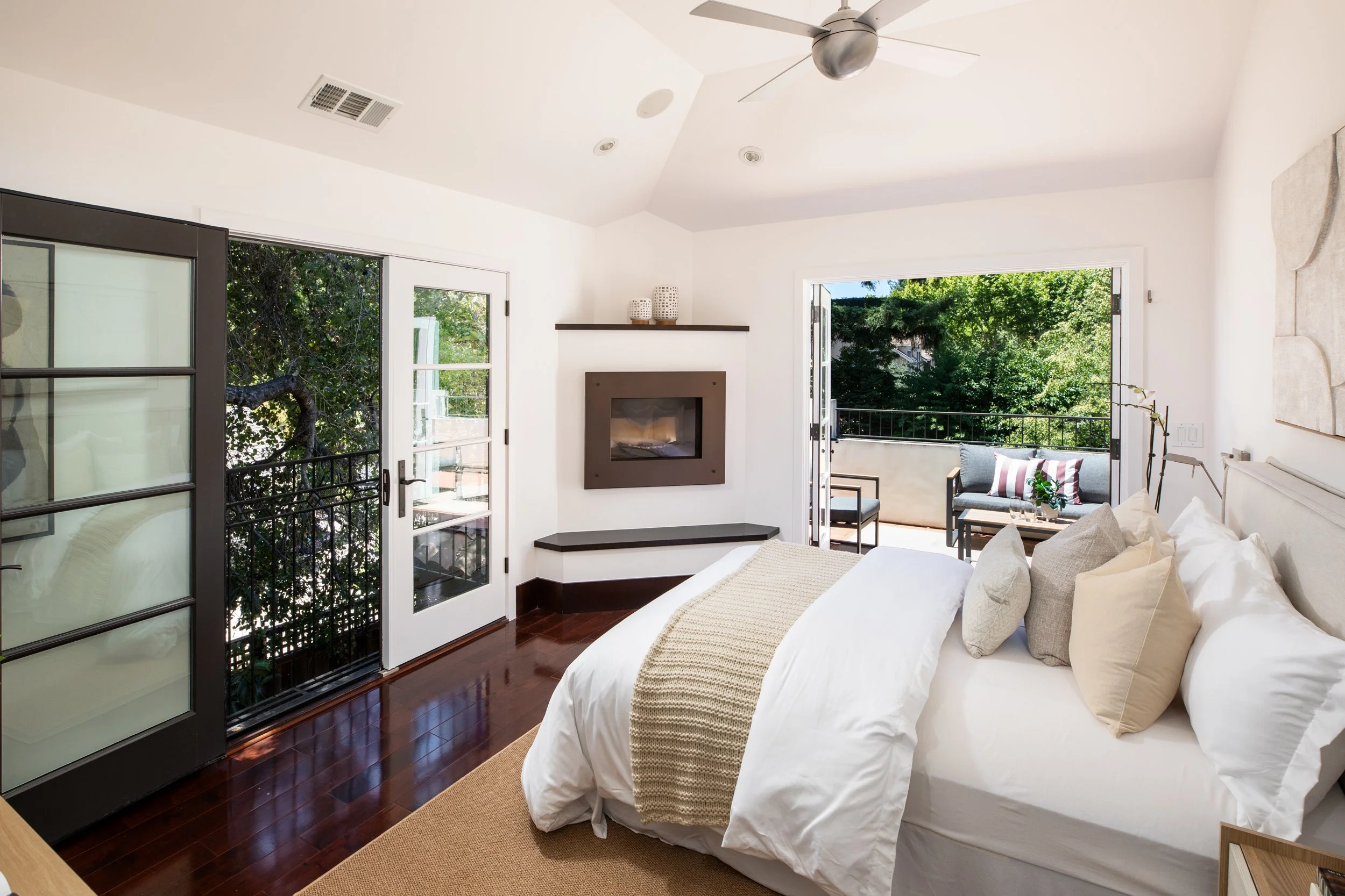A bright bedroom with white walls, a bed with white bedding and beige pillows, a wooden floor, large glass doors opening to a balcony with trees outside, and a ceiling fan.