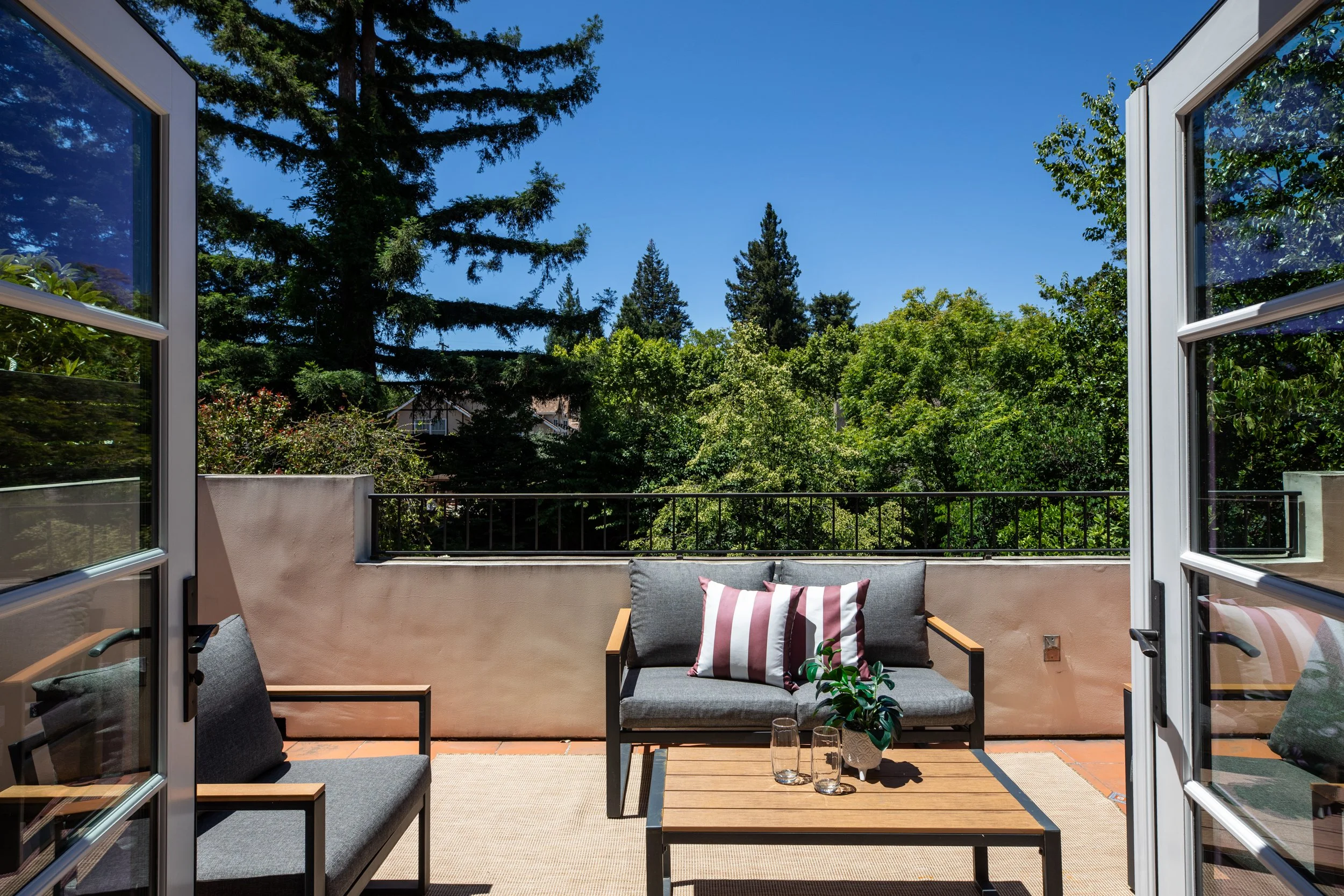 View from an open balcony door overlooking a lush green garden with tall trees and a clear blue sky.