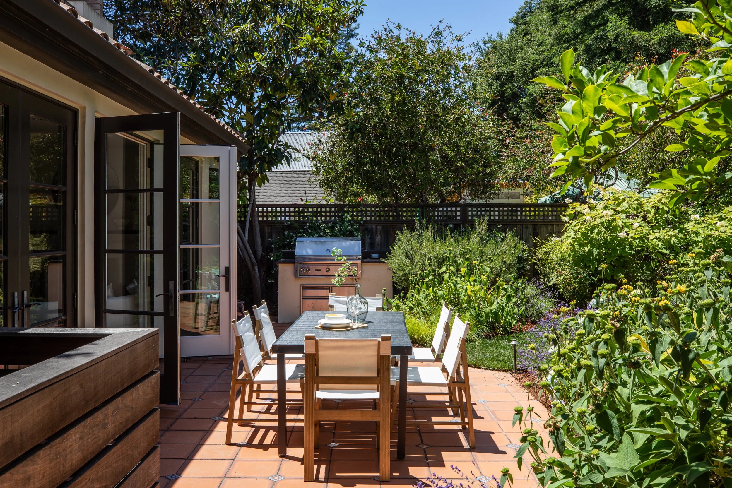 A backyard patio with a dining table set for a meal, surrounded by lush green trees and plants, with an open door leading to the house and an outdoor grill in the background.