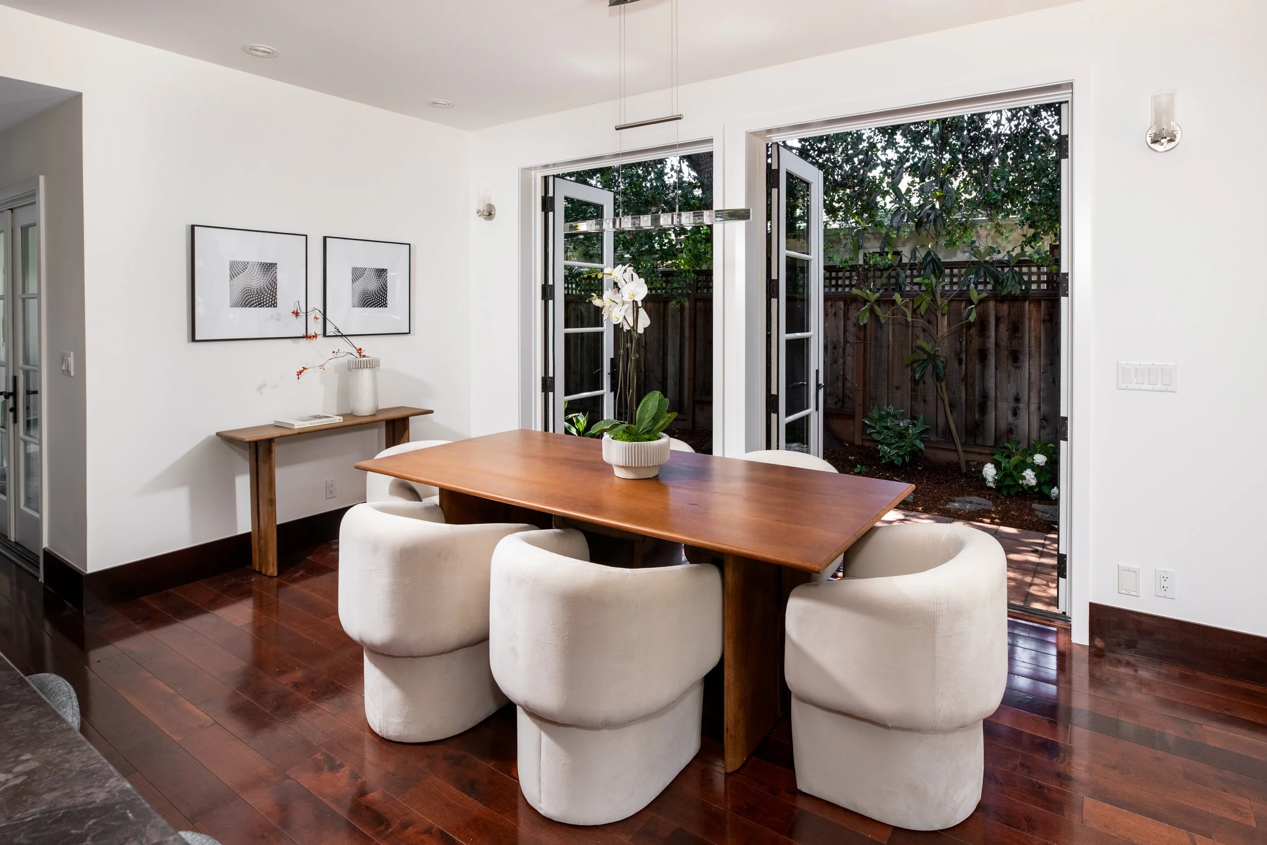 Dining area with a wooden table surrounded by six white upholstered chairs, view to outdoor yard with trees and wooden fence through open glass doors.