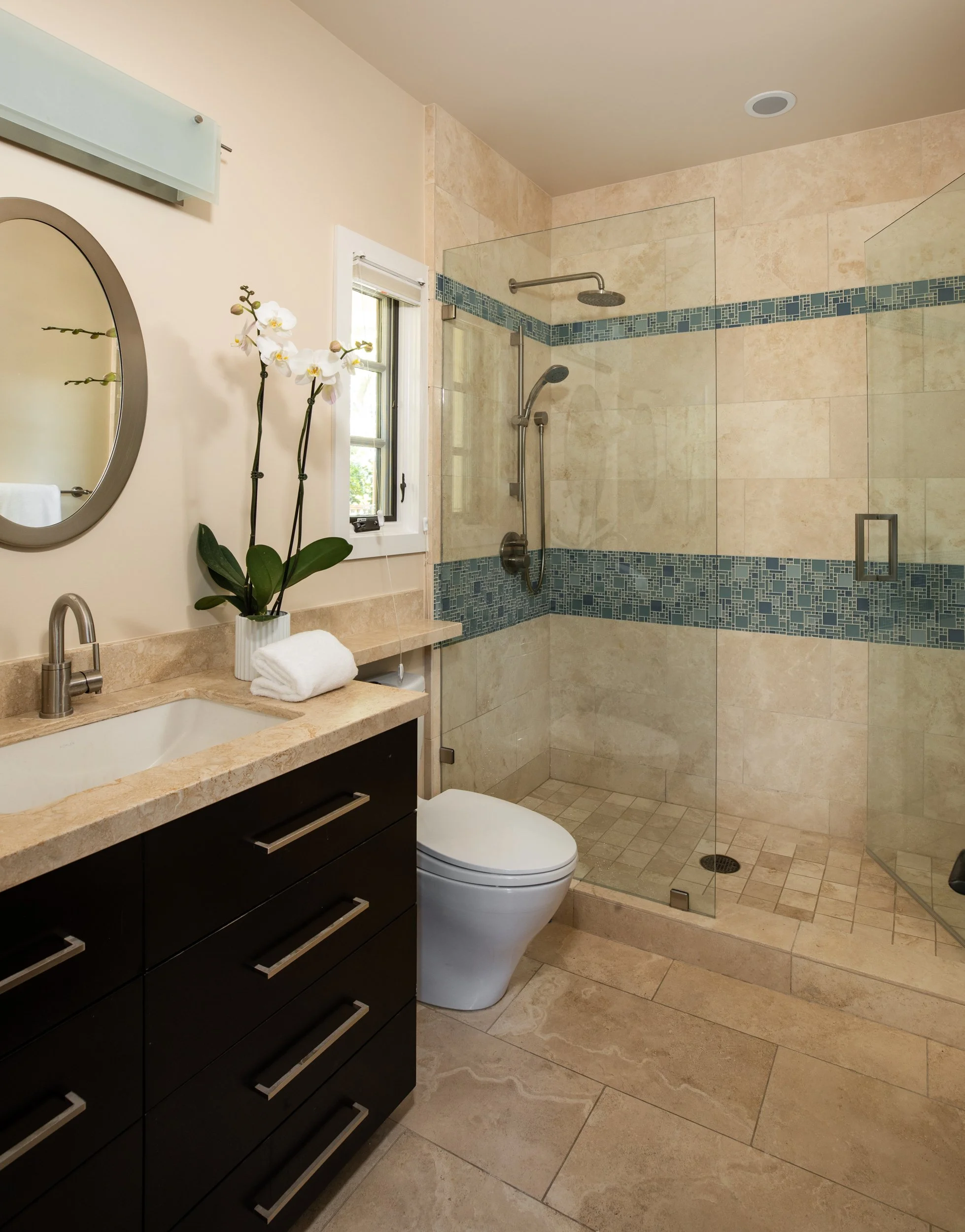A modern bathroom with a black vanity, beige granite countertop, and a white toilet. A round mirror hangs above the vanity, and a white orchid in a pot is on the counter next to folded towels. There is a small window with a white frame, and a glass-e