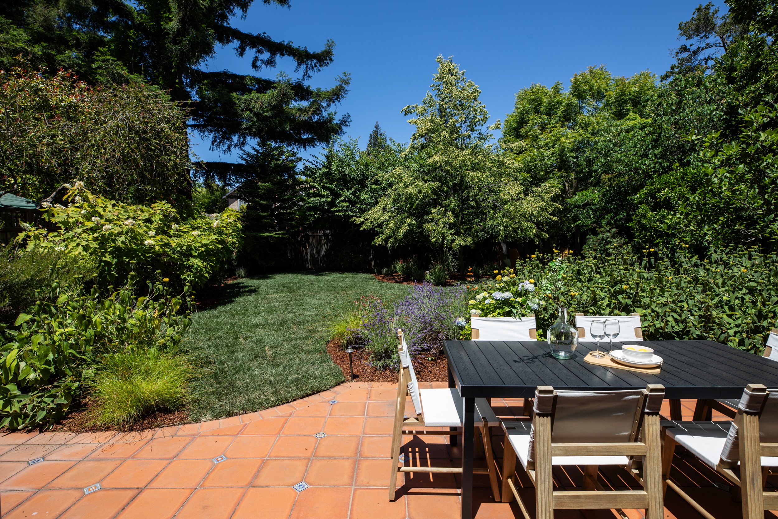 A backyard garden with a patio area featuring a black dining table set with plates, glasses, and a wine bottle, surrounded by lush greenery, flowering plants, and trees under a clear blue sky.