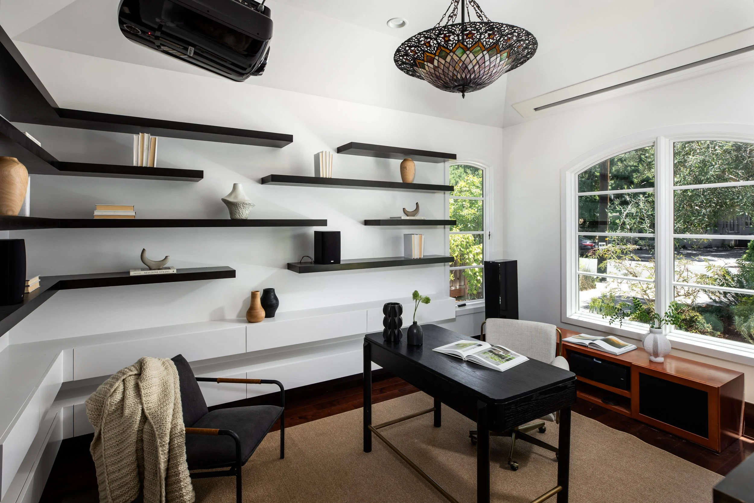 A modern living room with white walls and large windows showing greenery outside. Black floating shelves decorated with books and vases, a black coffee table with black vases and a plant, a gray chair with a beige knit blanket, and a colorful chandel