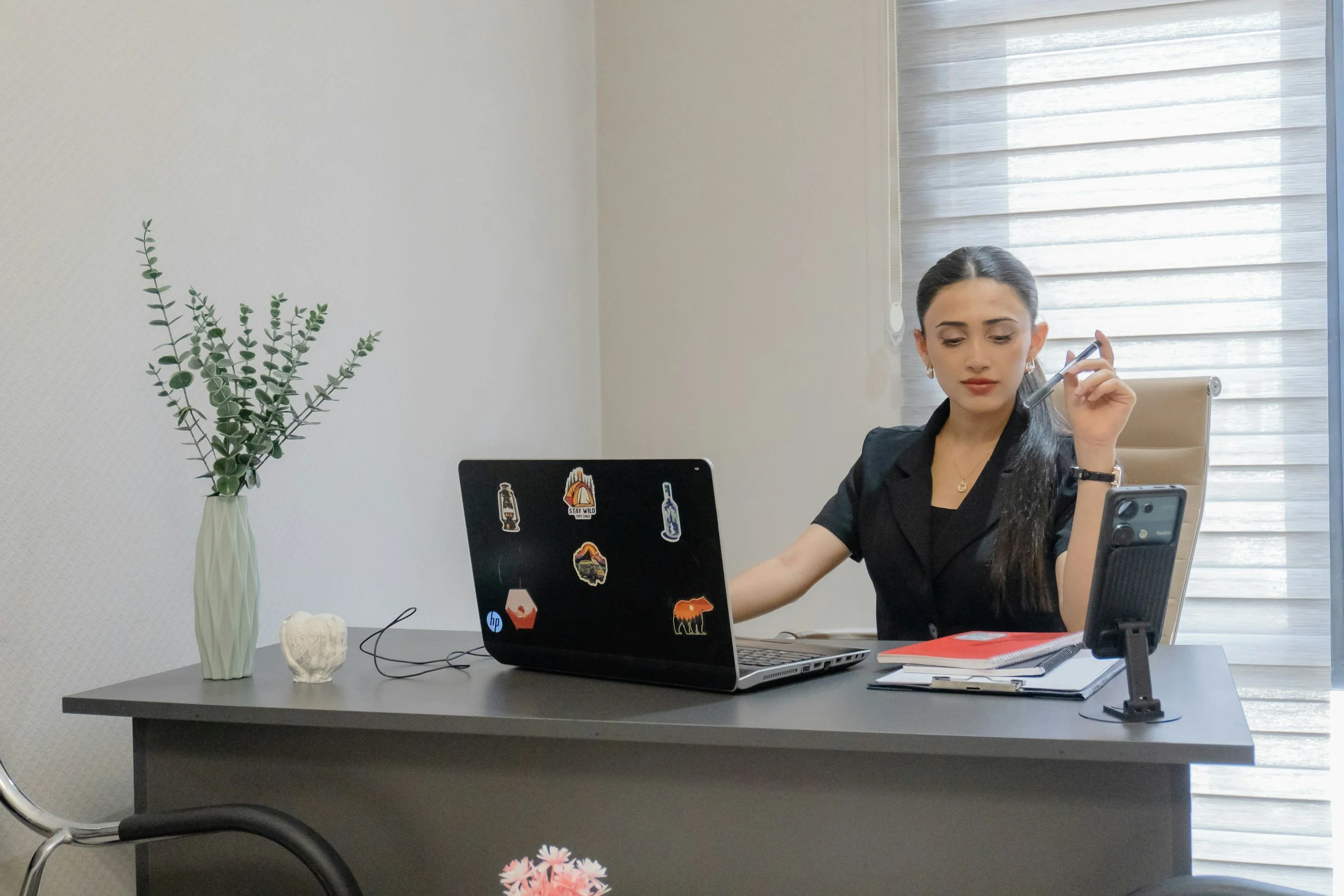 A woman with dark hair in a ponytail sitting at a desk with a laptop, holding a pen, in a well-lit room with sheer curtains. There is a large vase with green foliage, a small skull, and a smartphone on the desk.