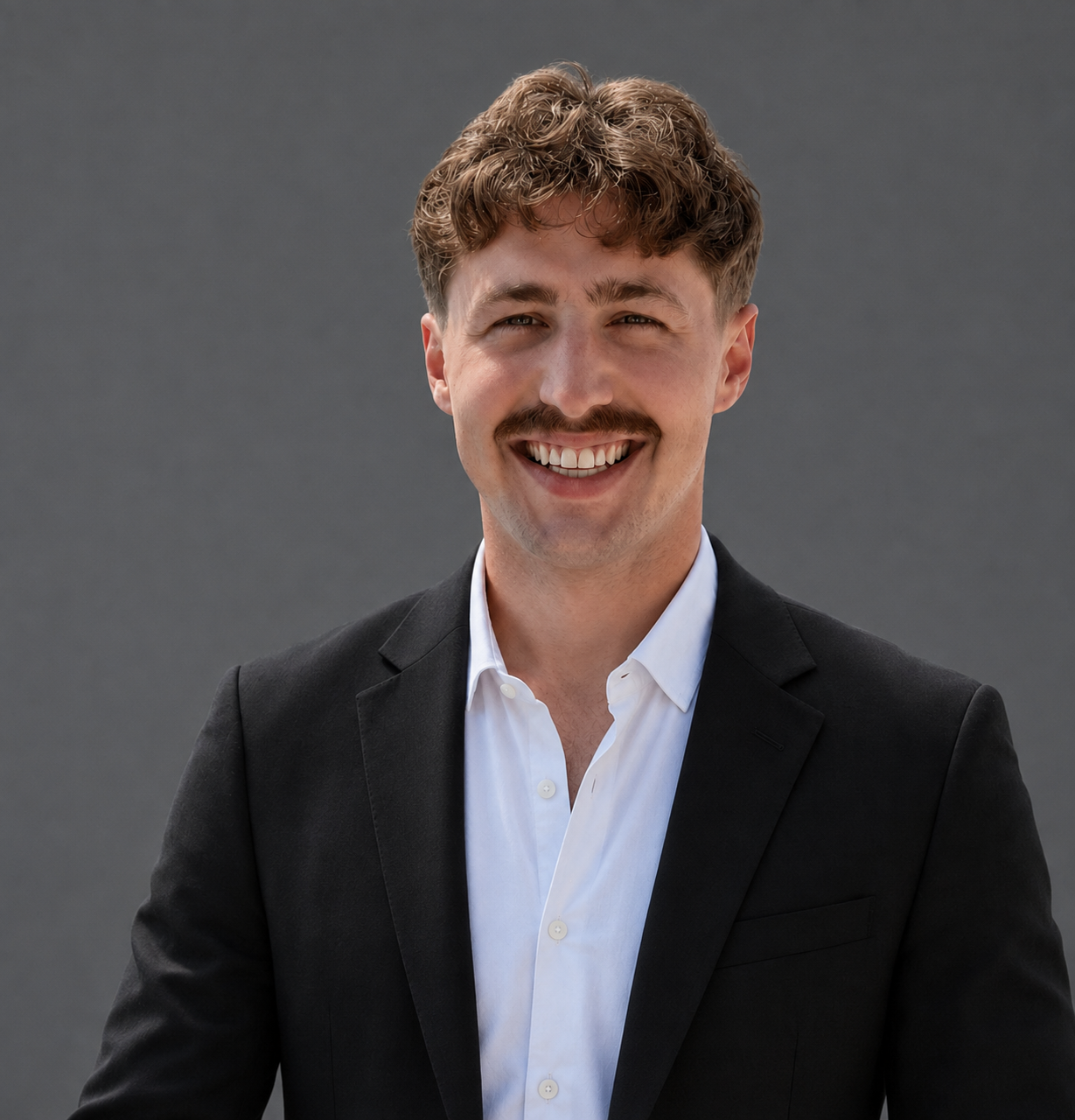 Professional headshot of the Senior Sales Manager at TG Talent Team LLC, featuring a young man with blonde hair, a mustache, and a broad smile, dressed in a black blazer and white shirt against a neutral gray background.