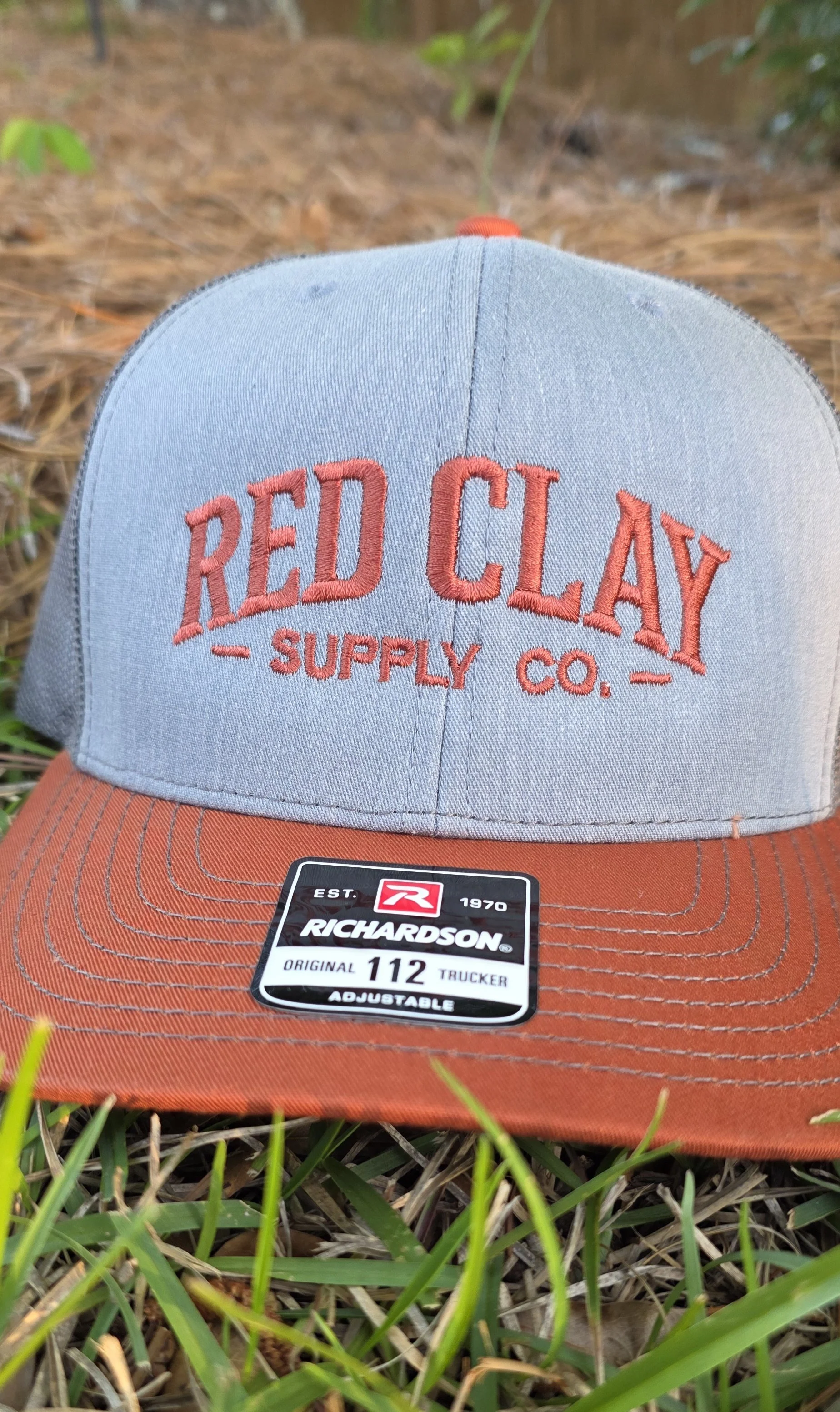 A baseball cap with the words 'Red Clay Supply Co.' embroidered on the front, placed on grass. The cap has a light gray crown with orange stitching and an orange bill. There is a sticker on the bill that says 'Richardson Original 112 Trucker Adjustable.'