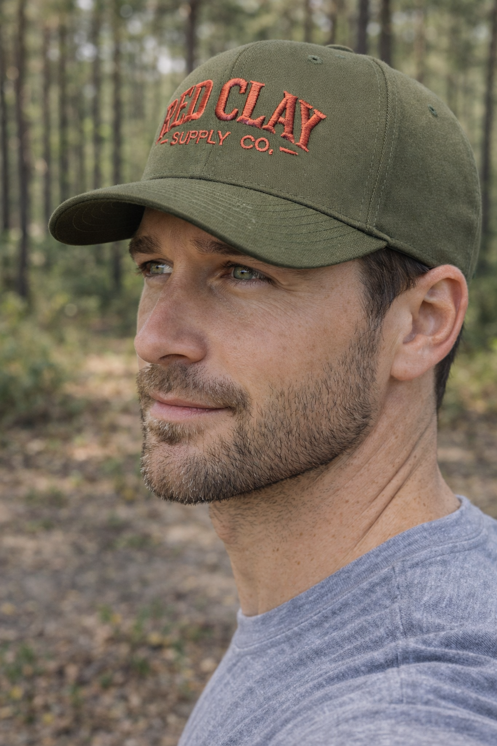 Close-up of a man wearing a green baseball cap with red embroidered text, standing outdoors in a forest with trees in the background.