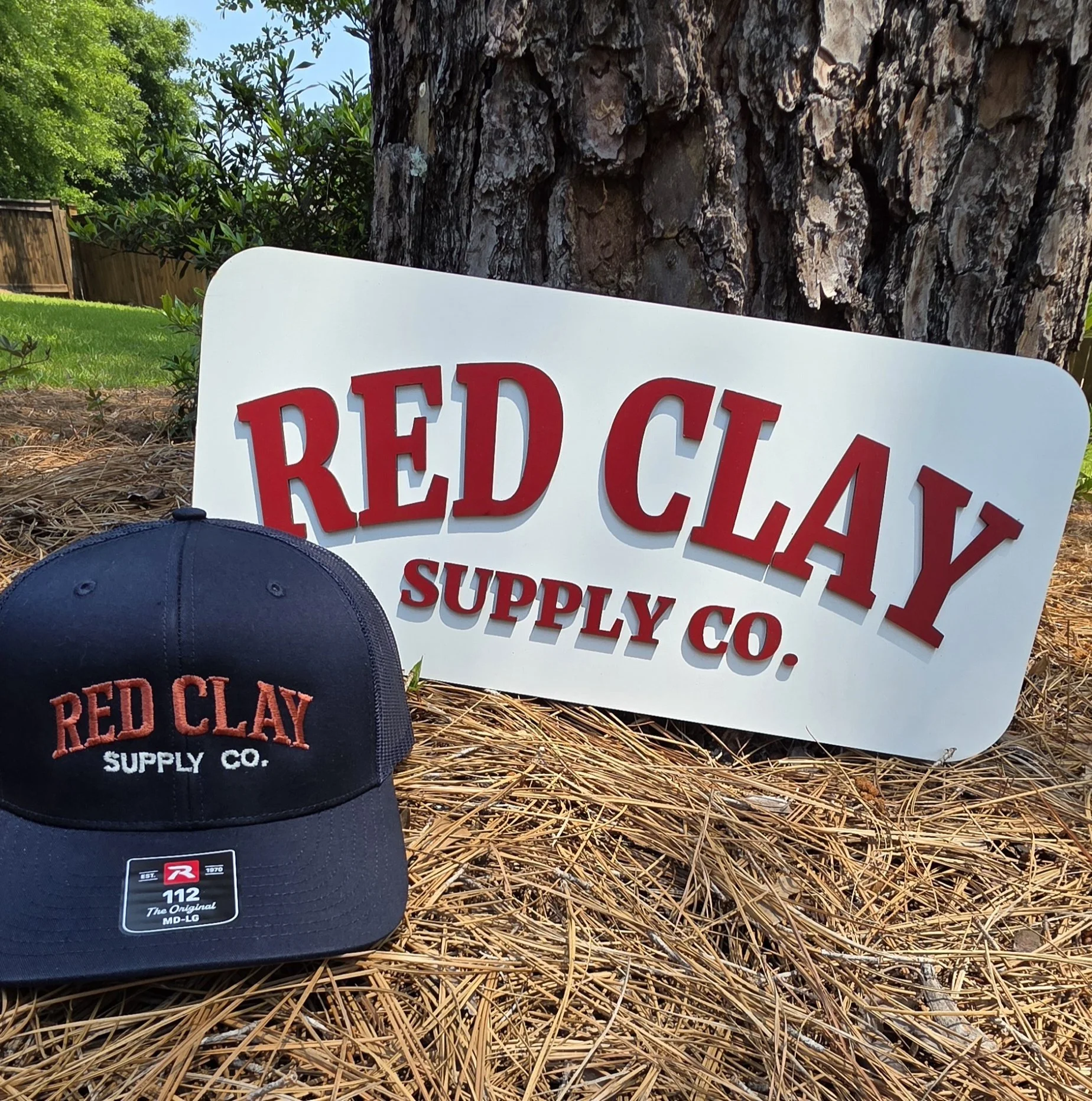 A black baseball cap with red and white text that reads 'Red Clay Supply Co.' placed on straw-like ground next to a white sign with red text saying the same. The background features a large tree trunk, green bushes, and a blue sky.