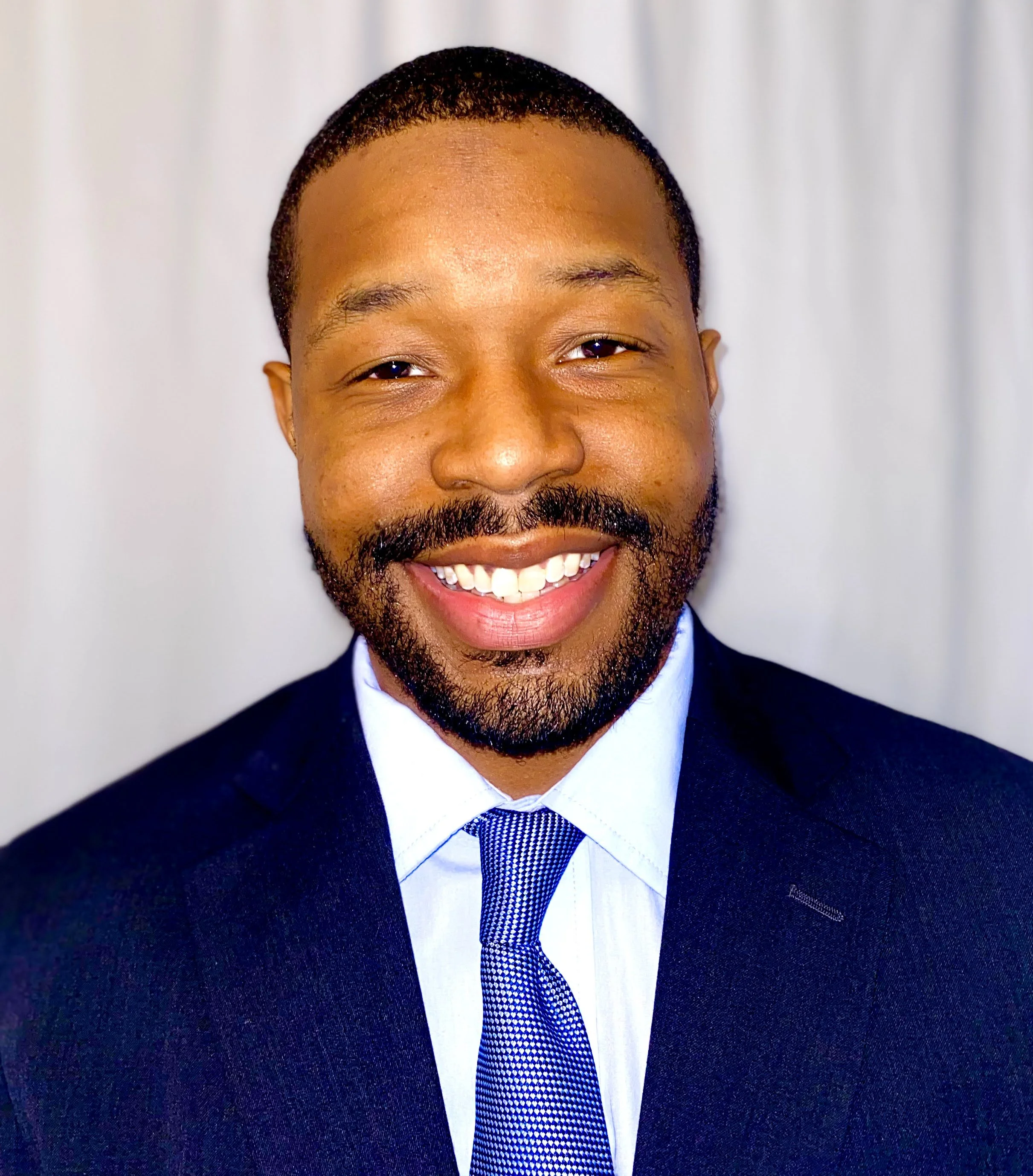 Close-up of a smiling man wearing a dark suit, white shirt, and blue tie with white patterns, standing against a light background.