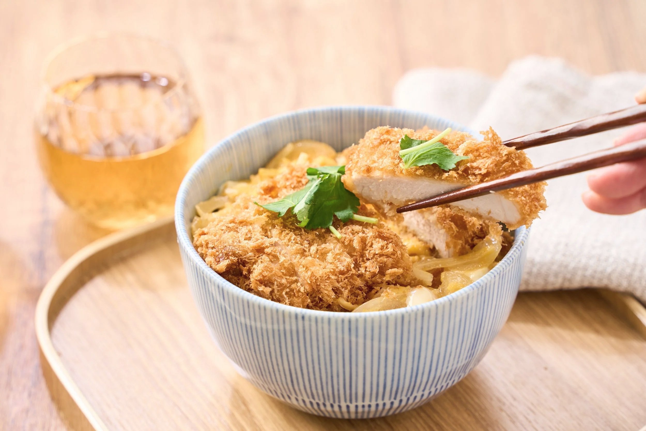A bowl of panko fried pork cutlet with herbs, served over noodles on a blue and white striped bowl. A pair of chopsticks holding a piece of pork. In the background, a glass of beer and a small dish, possibly of sauce, on a wooden tray.