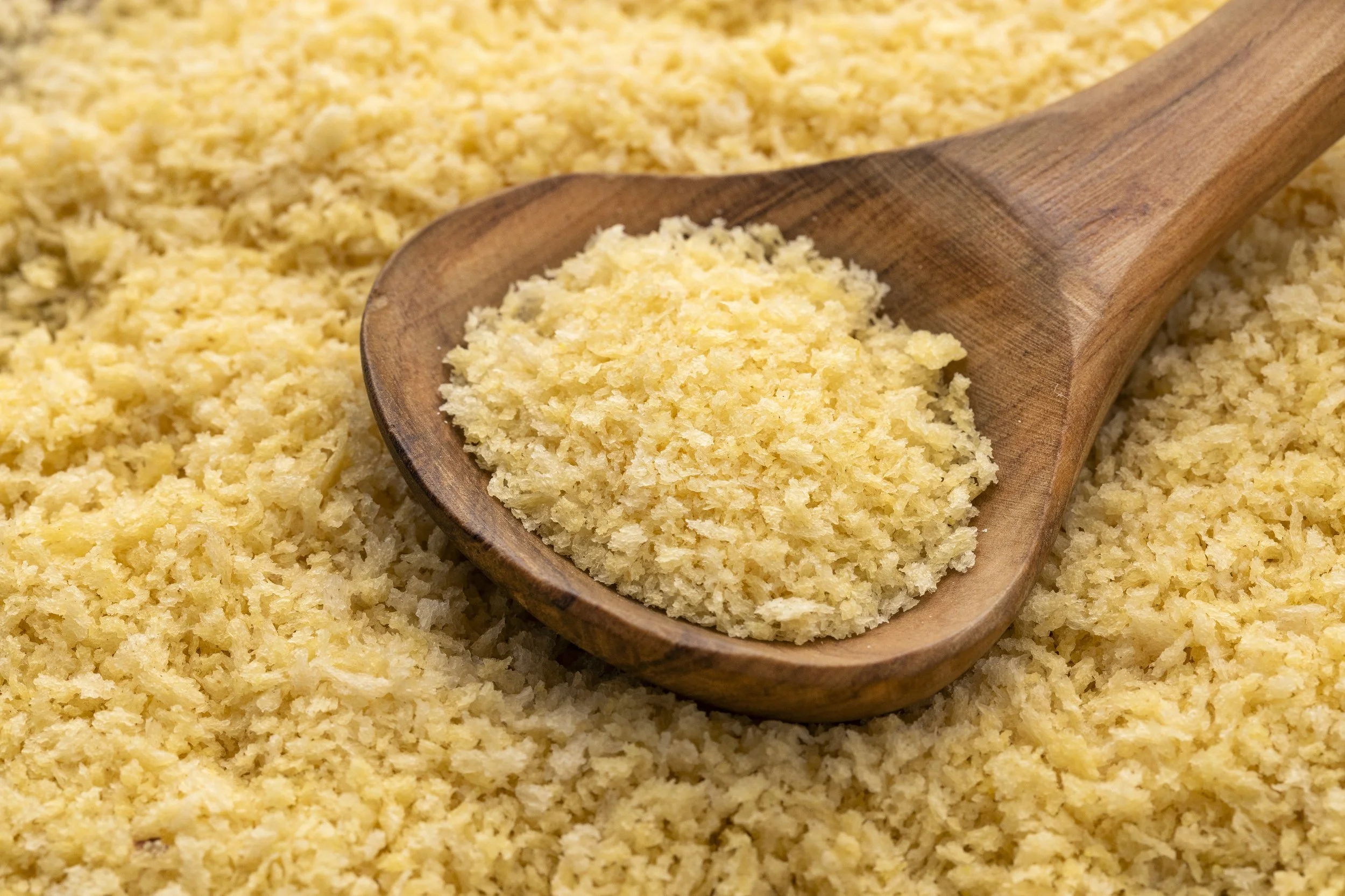 Close-up of a wooden scoop filled with yellow panko breadcrumbs, in a bowl of yellow panko breadcrumbs.