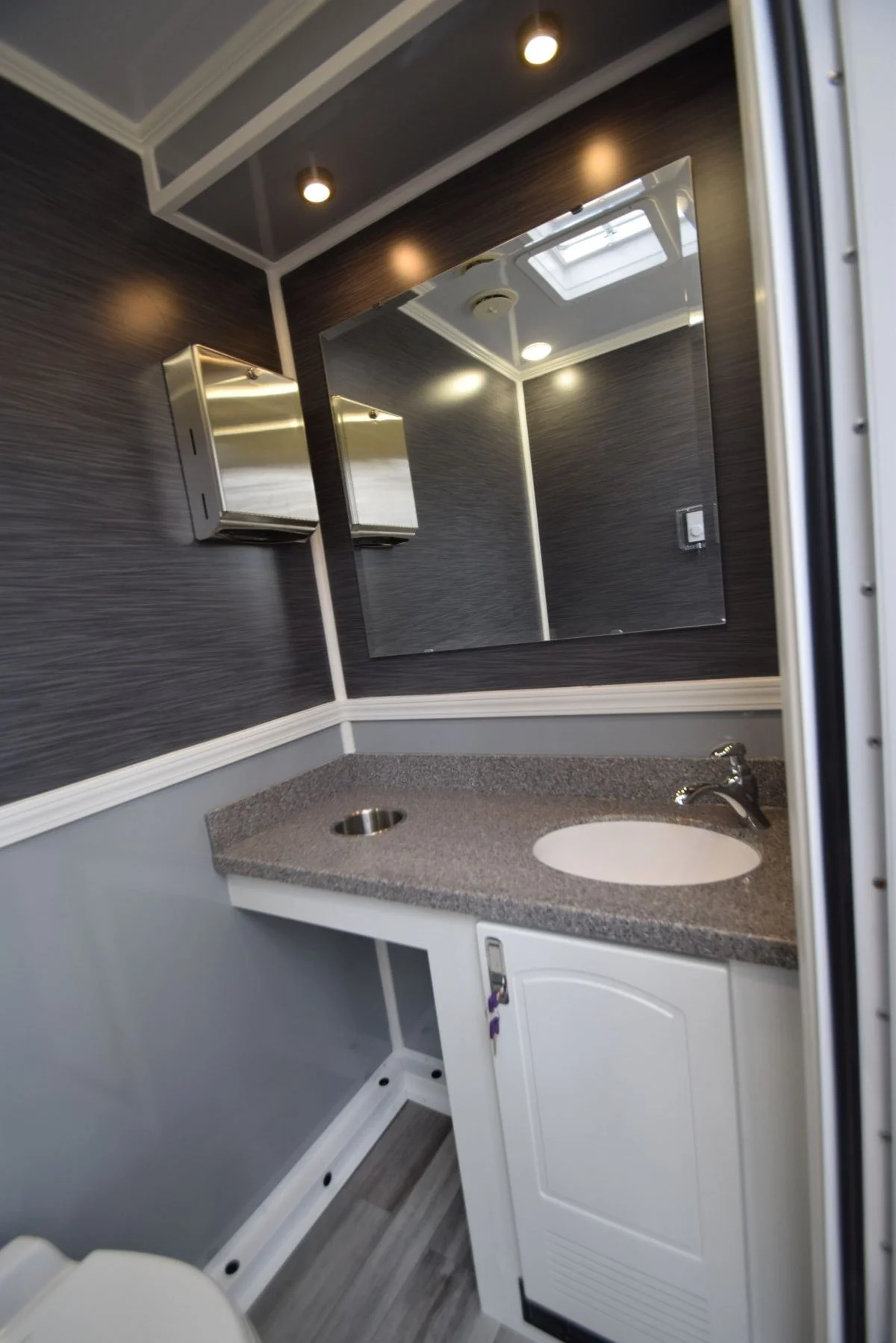 Small bathroom with a granite countertop, cabinet, large mirror, and wood textured walls; ceiling lights and skylight.