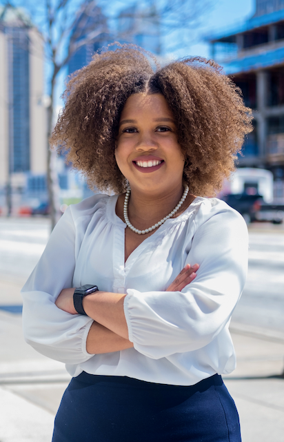A woman with curly hair, wearing a white blouse, pearl necklace, and a smartwatch, standing outdoors with her arms crossed and smiling.