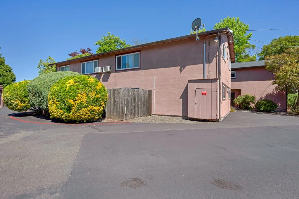 A two-story pink apartment building with a satellite dish on the roof, surrounded by green trees and bushes, and a paved parking area in front.