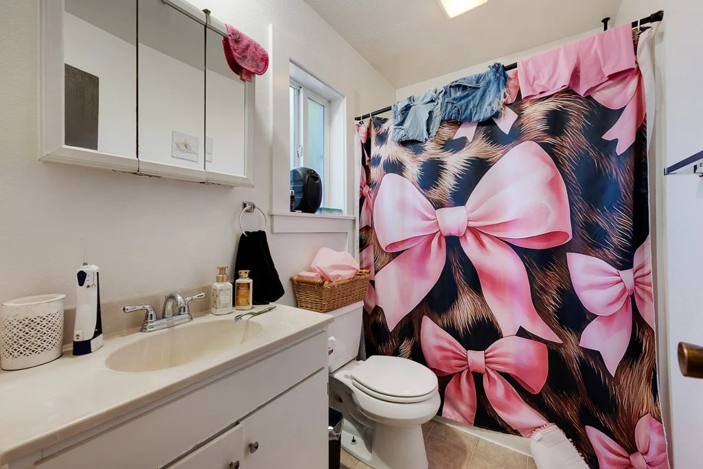 A small bathroom with a white sink, a mirror cabinet, and a toilet. There is a pink bow-patterned shower curtain hanging and clothes draped over the top. Items such as soap, lotion, and a basket are on the sink counter, and a small window is above th