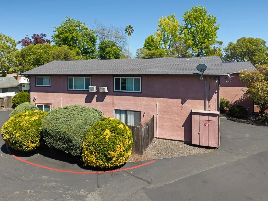 Pink two-story apartment building with a gray shingle roof, two window air conditioning units, a satellite dish, and a fenced backyard, surrounded by green trees and bushes, with a paved parking lot in front.