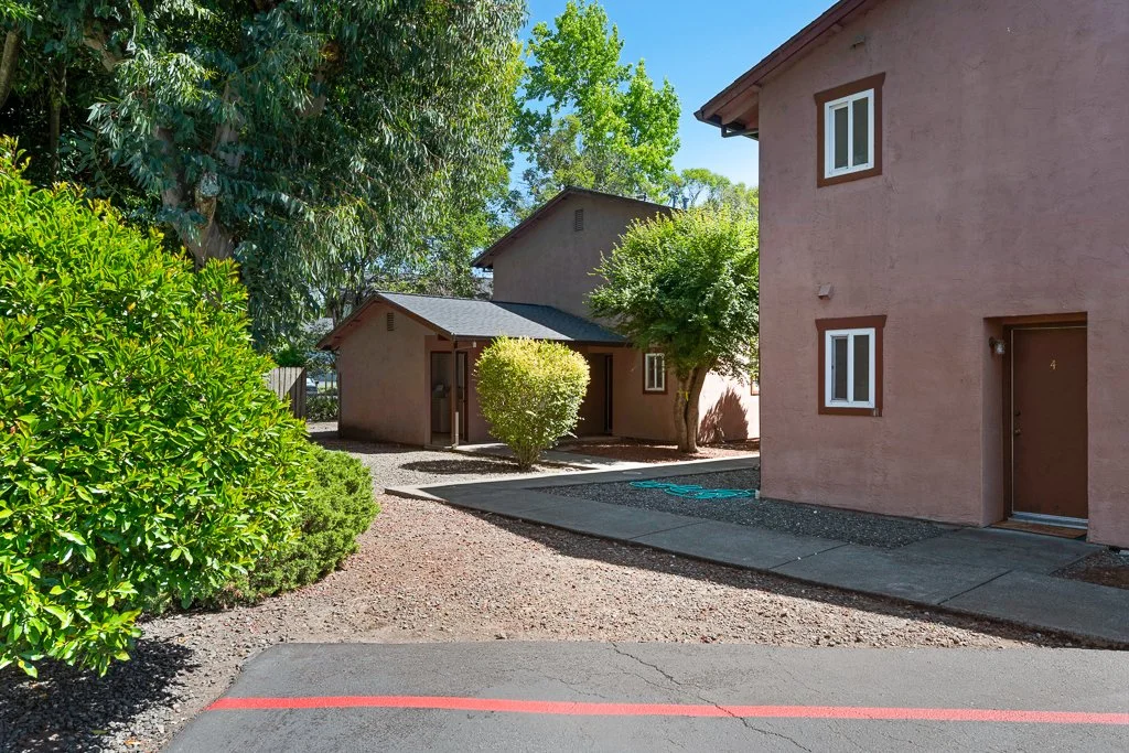 Exterior view of a two-story apartment complex with a pinkish stucco finish, surrounded by bushes and trees, with a gravel pathway and parking lot in the foreground.