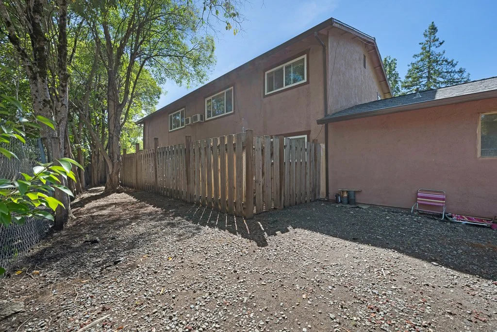 Backyard with dirt ground, surrounded by a wooden fence and trees, with a two-story house in the background.
