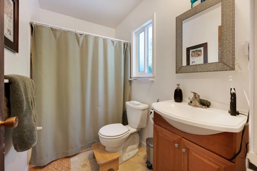 Small bathroom with a toilet next to a window, a wooden step stool, a beige shower curtain, and a corner sink with a mirror above it.