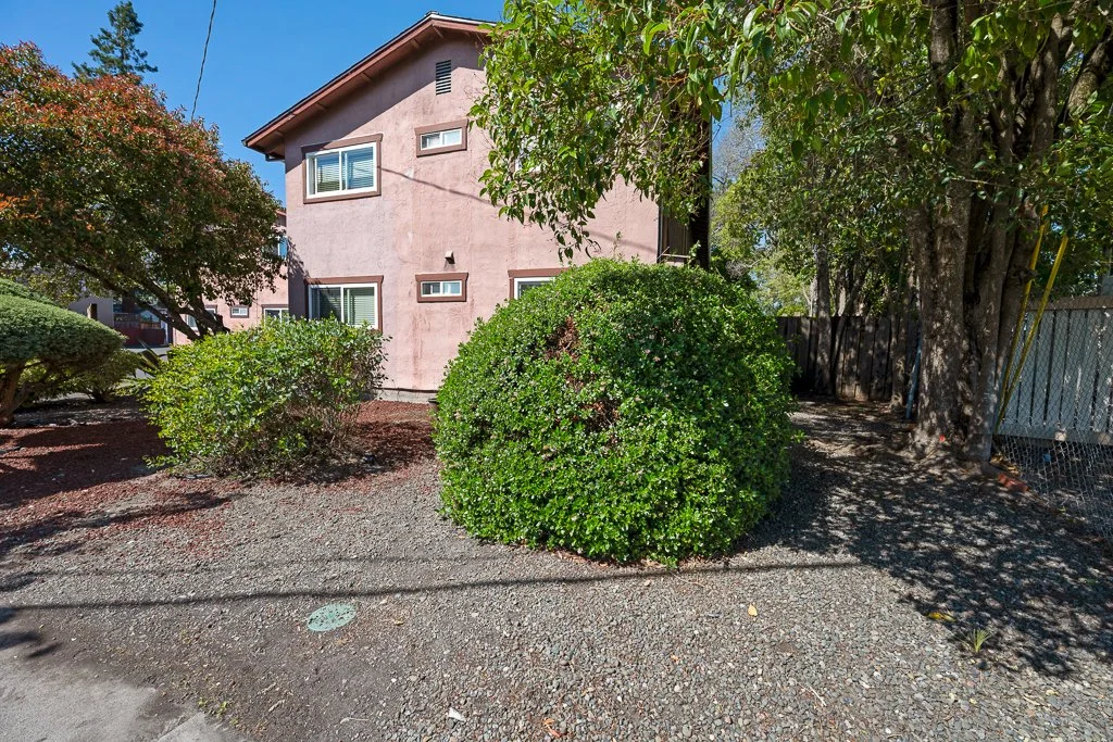 A pink two-story house with small rectangular windows, surrounded by green bushes and trees, on a sunny day.