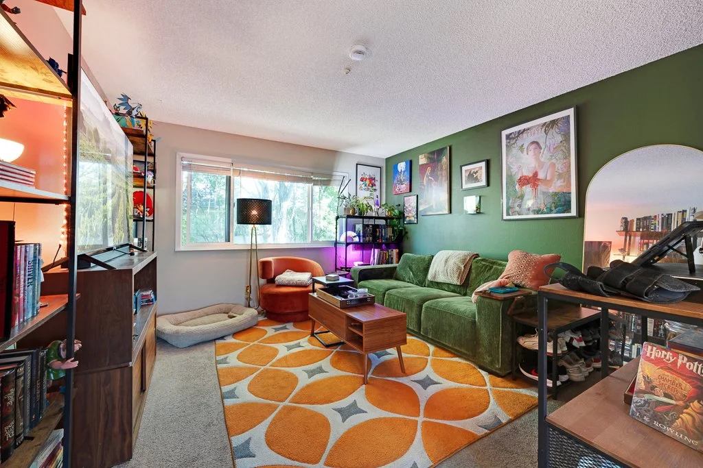 Living room with green sofa, orange armchair, bookshelves, artwork on green wall, window with blinds, rug with orange and gray pattern, and various decorations.
