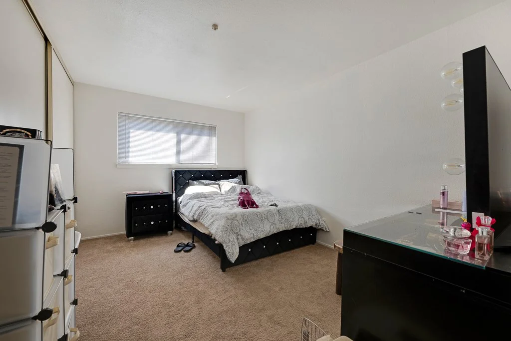 A bedroom with a bed, nightstand, dresser with mirror, and closet with sliding doors, beige carpet, white walls, window with blinds, and some personal items on the dresser.