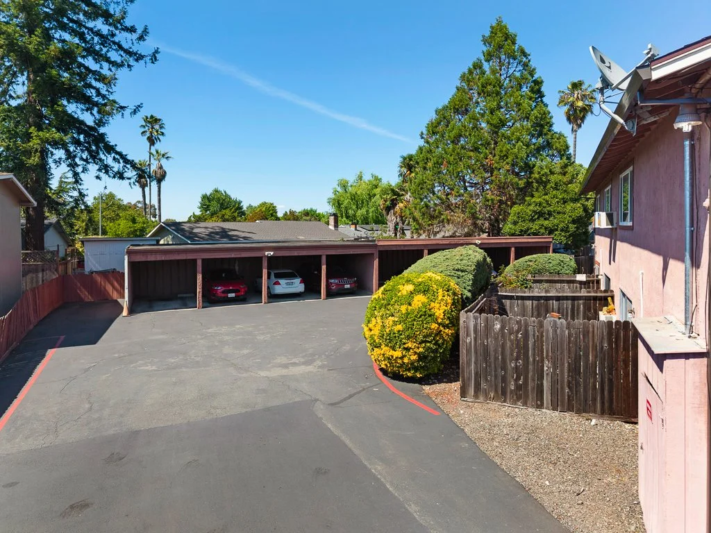 A small parking lot with three cars parked under a wooden structure, surrounded by residential buildings and trees, with a clear blue sky overhead.