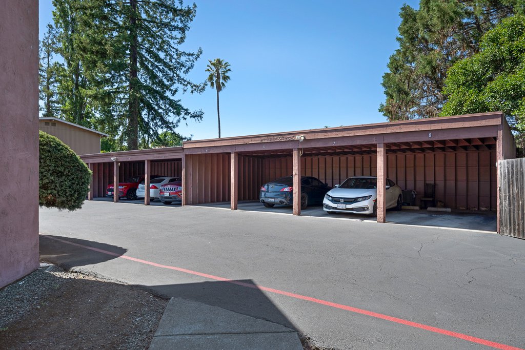 Apartment carport with five parked cars under a wooden structure, with trees in the background and a clear blue sky.