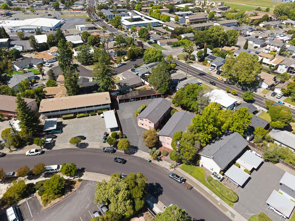 Aerial view of a suburban neighborhood with houses, trees, and streets, featuring a curved road with parked cars and a parking lot surrounded by residential buildings.
