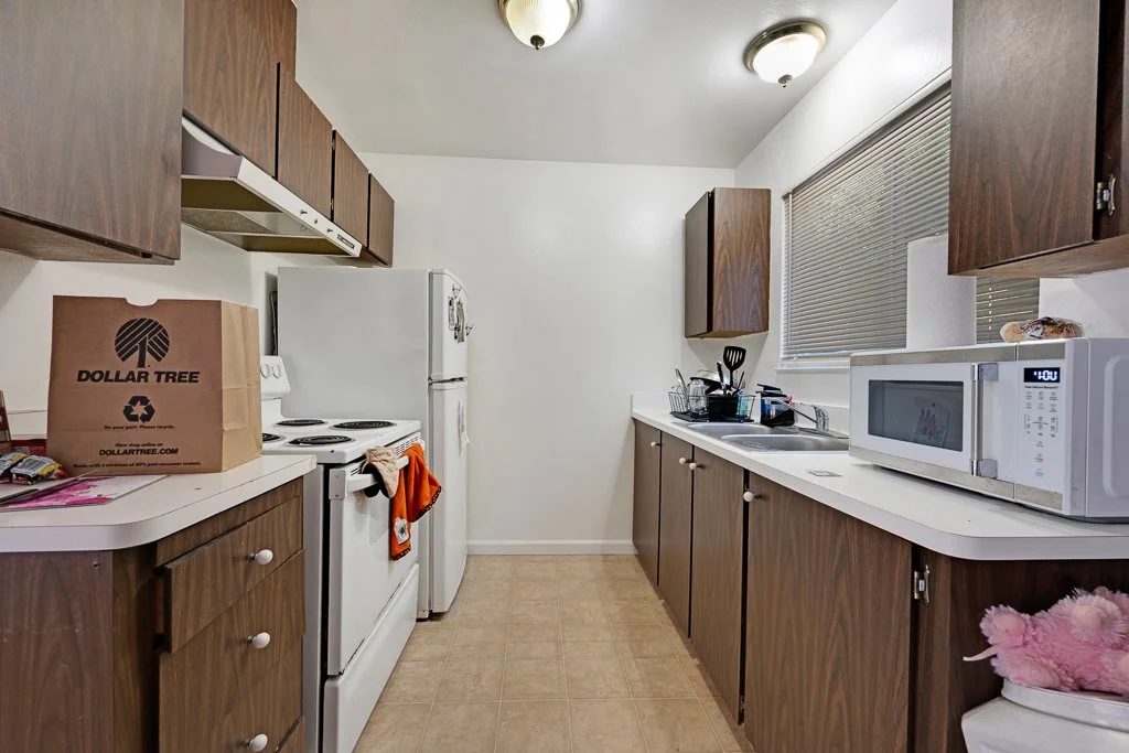 A small kitchen with wooden cabinets, white appliances including a refrigerator, stove, and microwave. There is a paper bag from Dollar Tree on the counter and various kitchen utensils near the sink.
