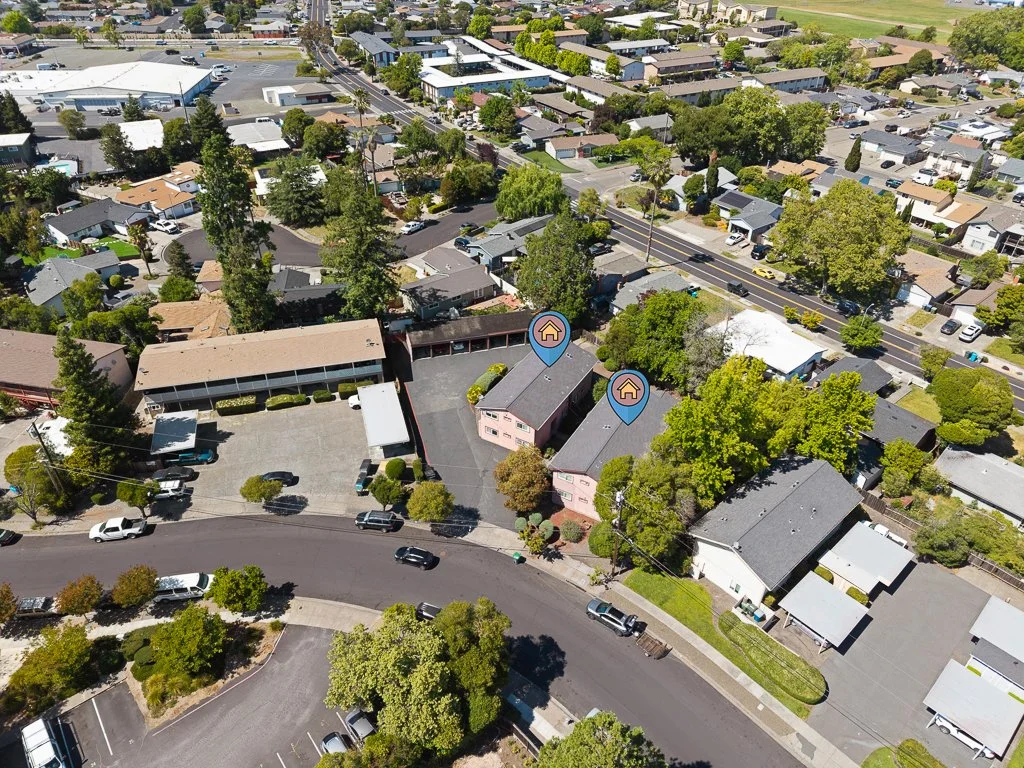 An aerial view of a residential neighborhood with houses, trees, and cars. Two pink-colored apartment buildings are marked with icons. A main road runs through the area.