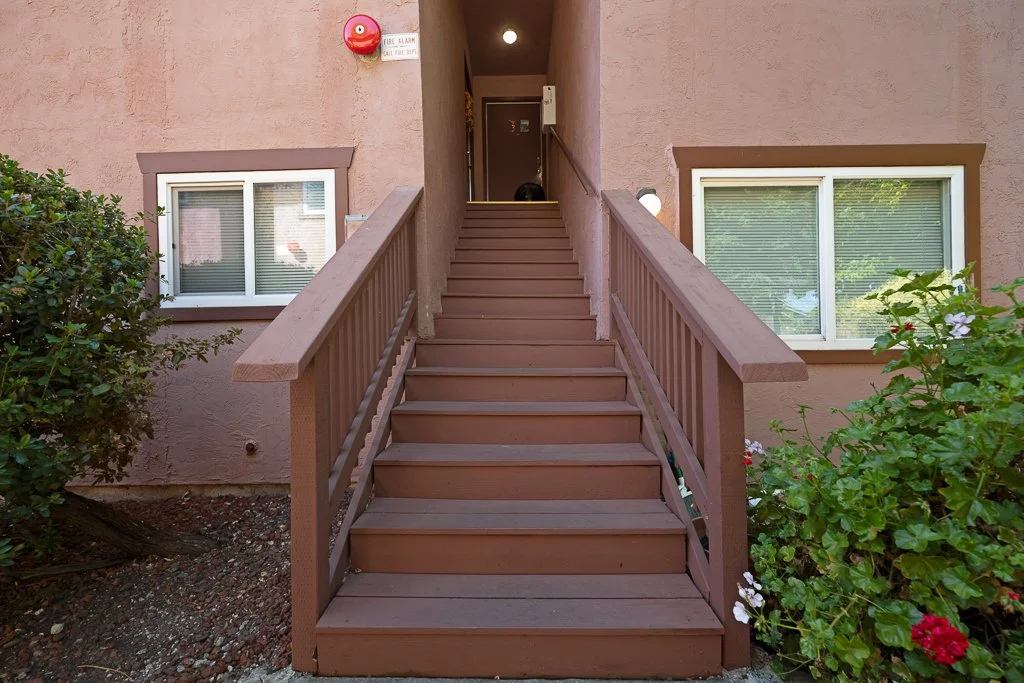 A wooden staircase leading up to a building entrance with pink walls, two windows, and surrounding green bushes and flowers.