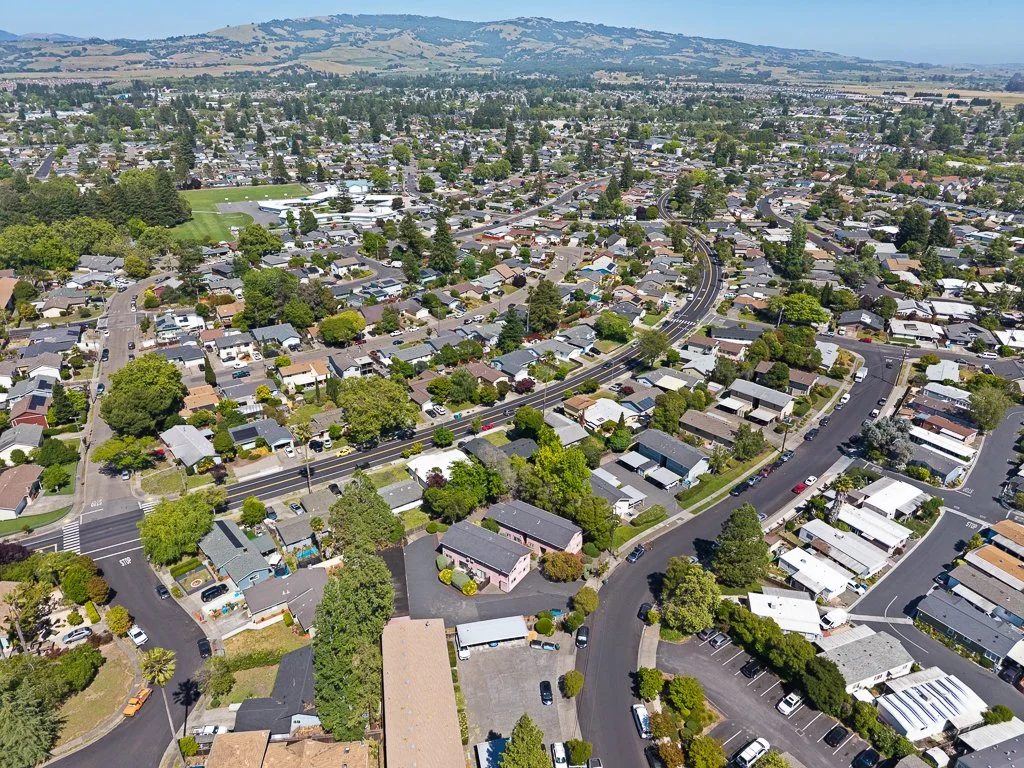 Aerial view of a suburban neighborhood with houses, trees, roads, and a sports field in the background, with hills in the distance.
