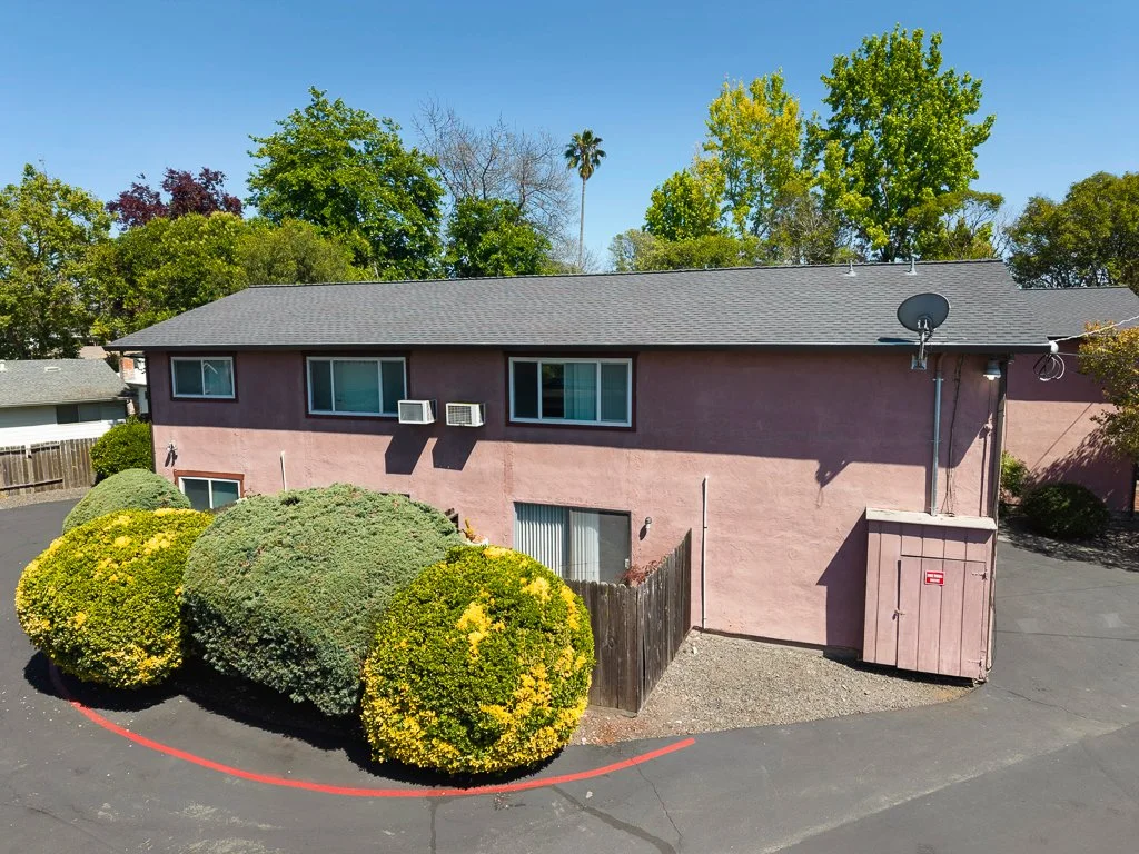Pink two-story apartment building with three bushes and trees in the background, satellite dish, and small fenced area outside.