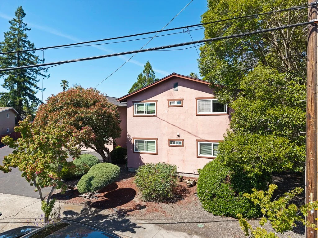 Two-story pink house with multiple windows, surrounded by trees and bushes, under a clear blue sky.