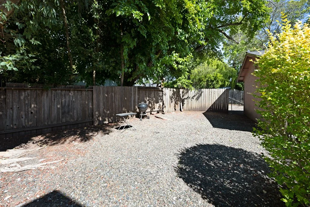 A backyard with a gravel ground, wooden fence, and trees providing shade. There is a small charcoal grill and a white folding chair near the fence. A shed is on the right side of the image, and a gate can be seen at the back.