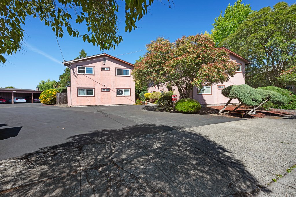 Two pink residential buildings surrounded by green trees and landscaped bushes, with a parking lot in front and a clear blue sky overhead.