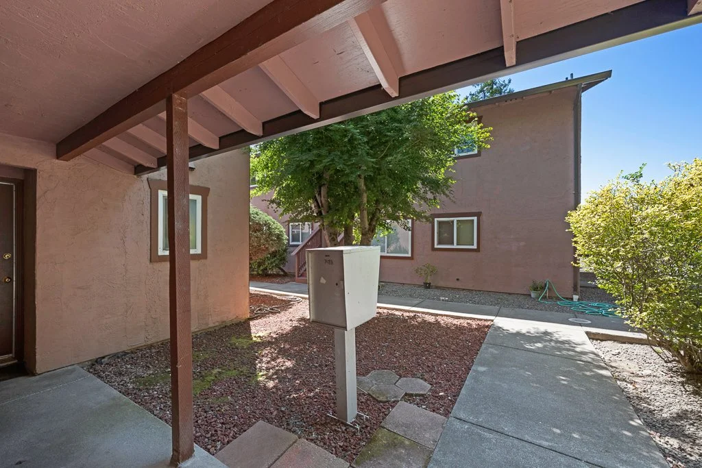 Apartment building exterior with walkway, tree in a pot, bushes, and sunlight.