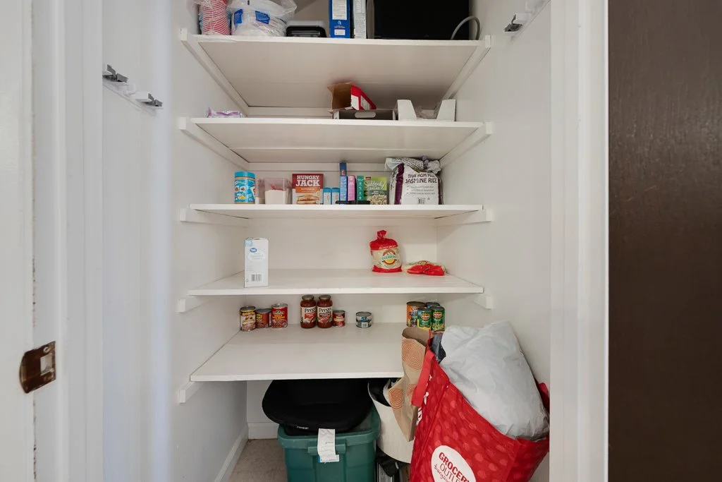 Empty pantry with a few canned goods, boxed food, and a bag of rice on the shelves, and a grocery sack with paper towels and a bag of rice on the floor.