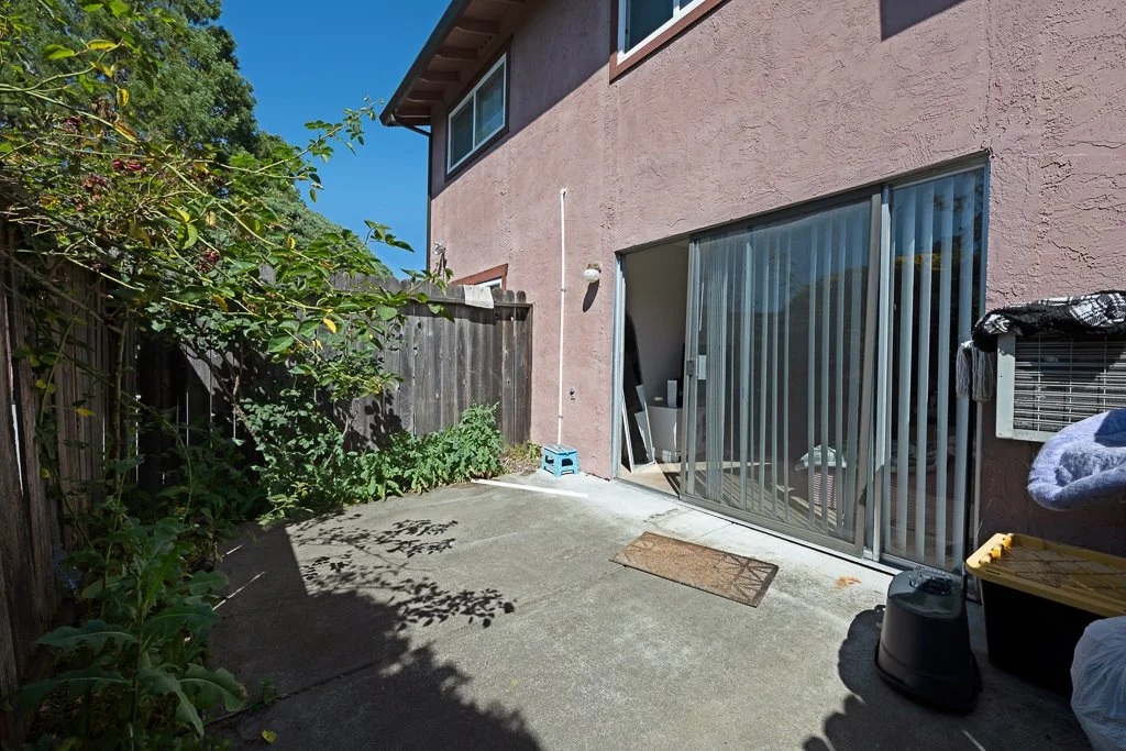 Small backyard patio area with concrete flooring, a sliding glass door, and a pink stucco house wall. There are plants along the left fence, and various outdoor items such as a trash can, a box, and a covered object on the right.