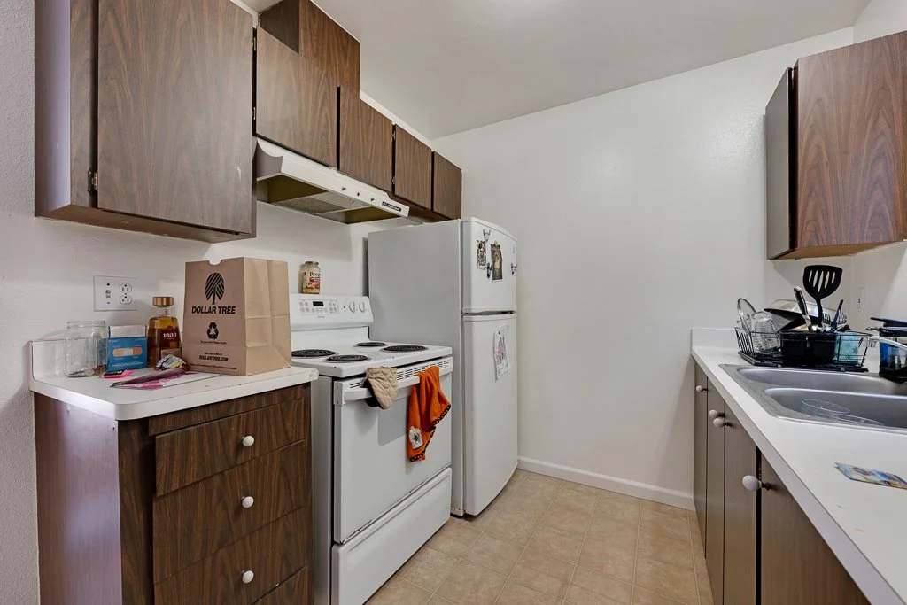 Small kitchen with white appliances, including a stove and refrigerator, and wooden cabinets. Countertop has a paper bag, jars, and eating utensils. The floor has beige tiles.