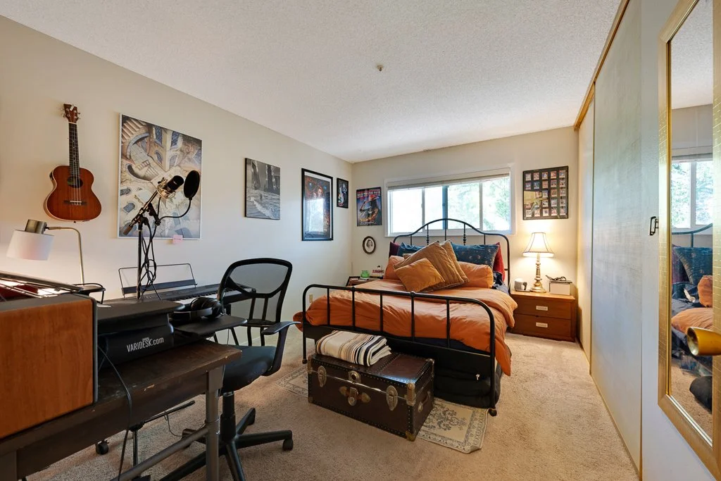 A bedroom with a bed, nightstand, lamp, and window, with wall art, a guitar, and a computer setup.