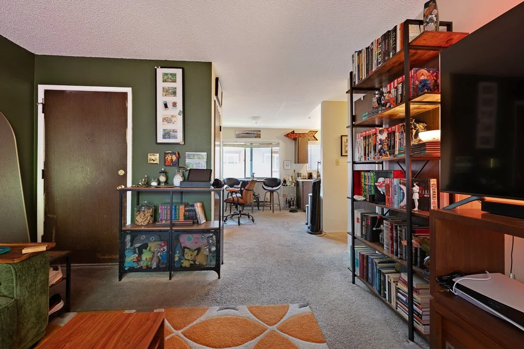 A view of a living room with a green wall, a dark door, and a bookshelf filled with books, with a television on the right side. The room extends into a dining area with a table and chairs, with natural light coming from windows in the background.